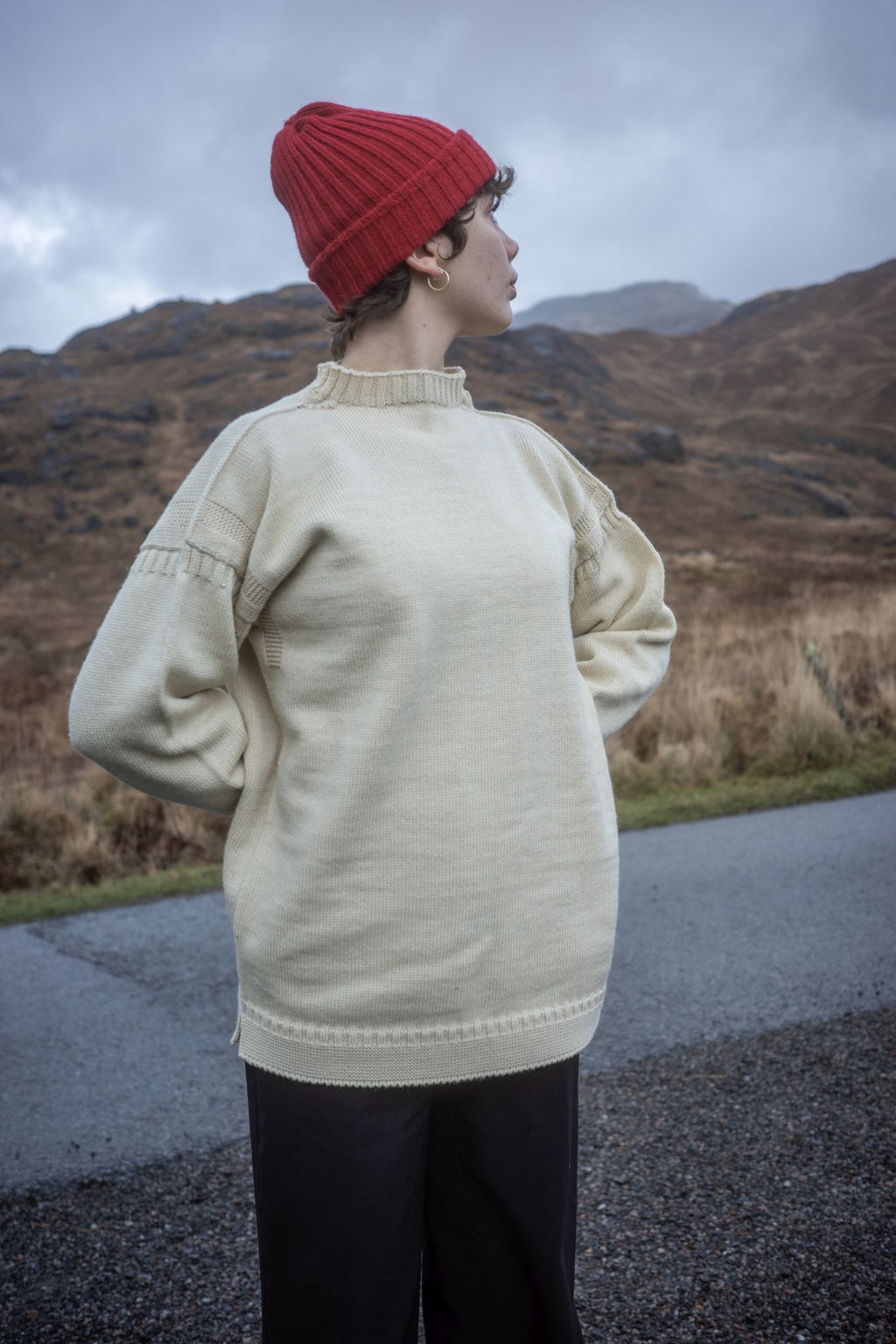 Female model wearing a cream coloured traditional Guernsey and  Red British Woollens Guernsey beanie while walking in mountains 