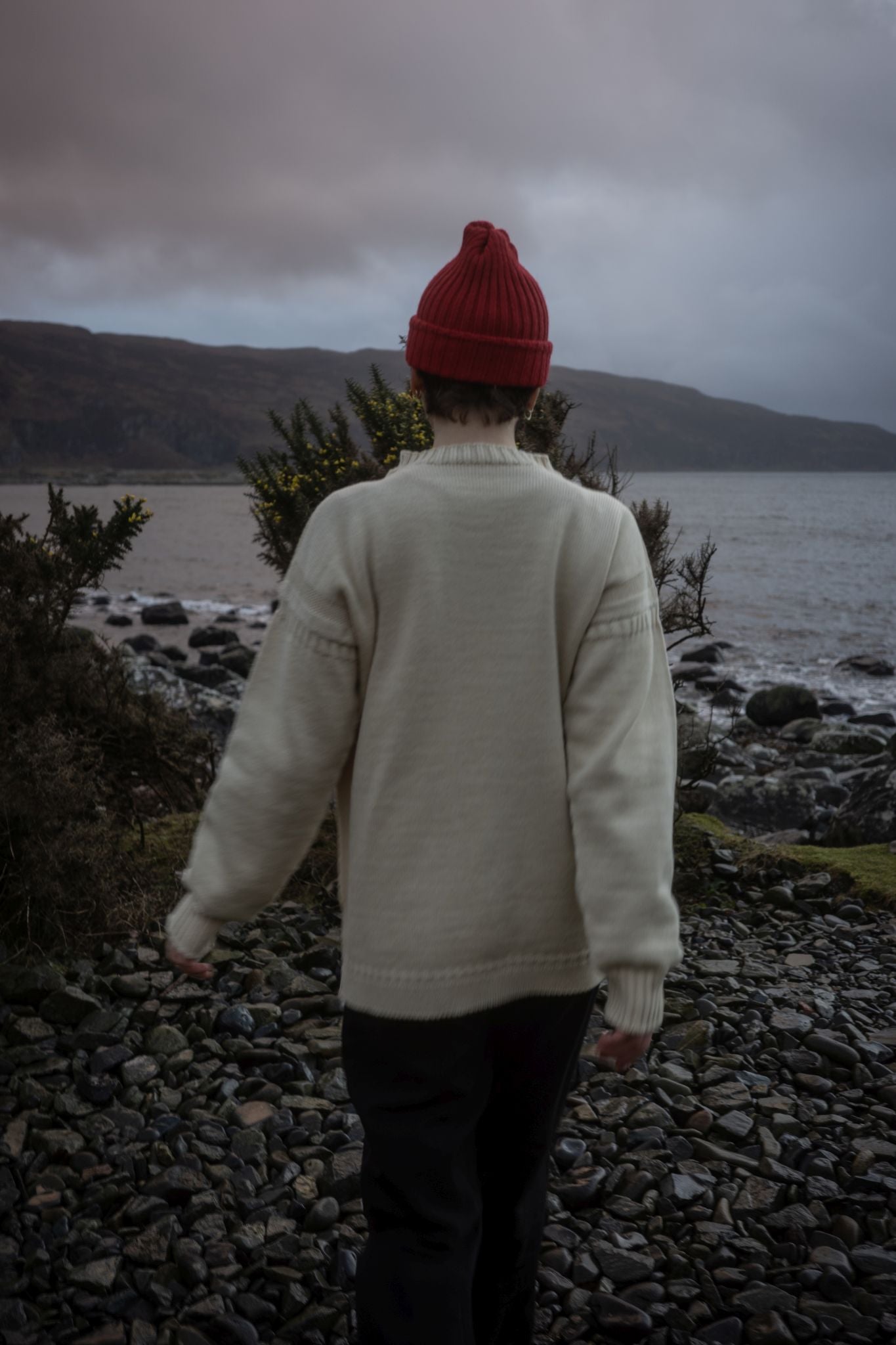Female model wearing a cream coloured traditional Guernsey and  Red British Woollens Guernsey beanie amongst gorse beside a lake with mountains behind 