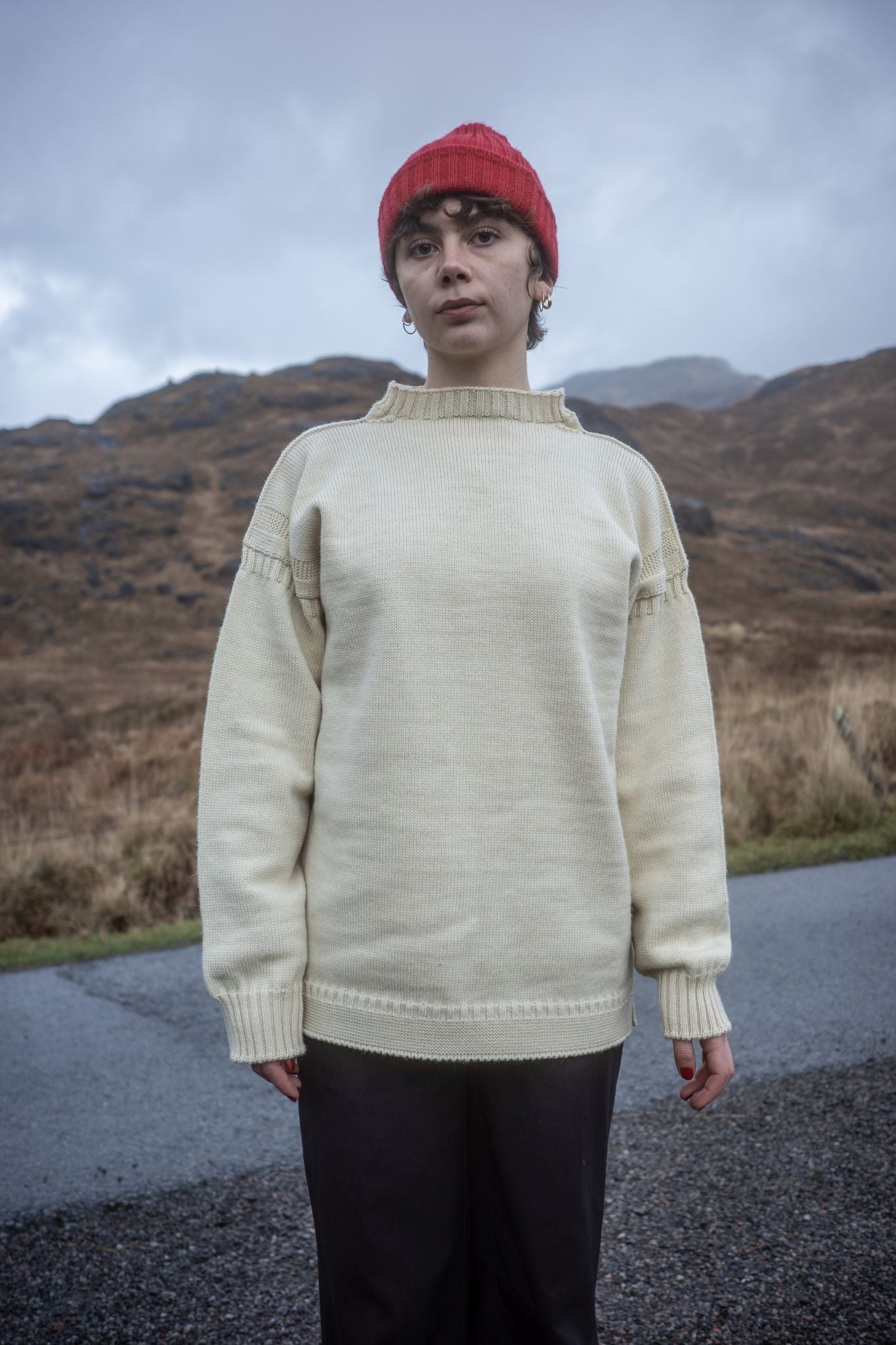Female model wearing a cream coloured traditional Guernsey and  Red British Woollens Guernsey beanie on a mountain road 