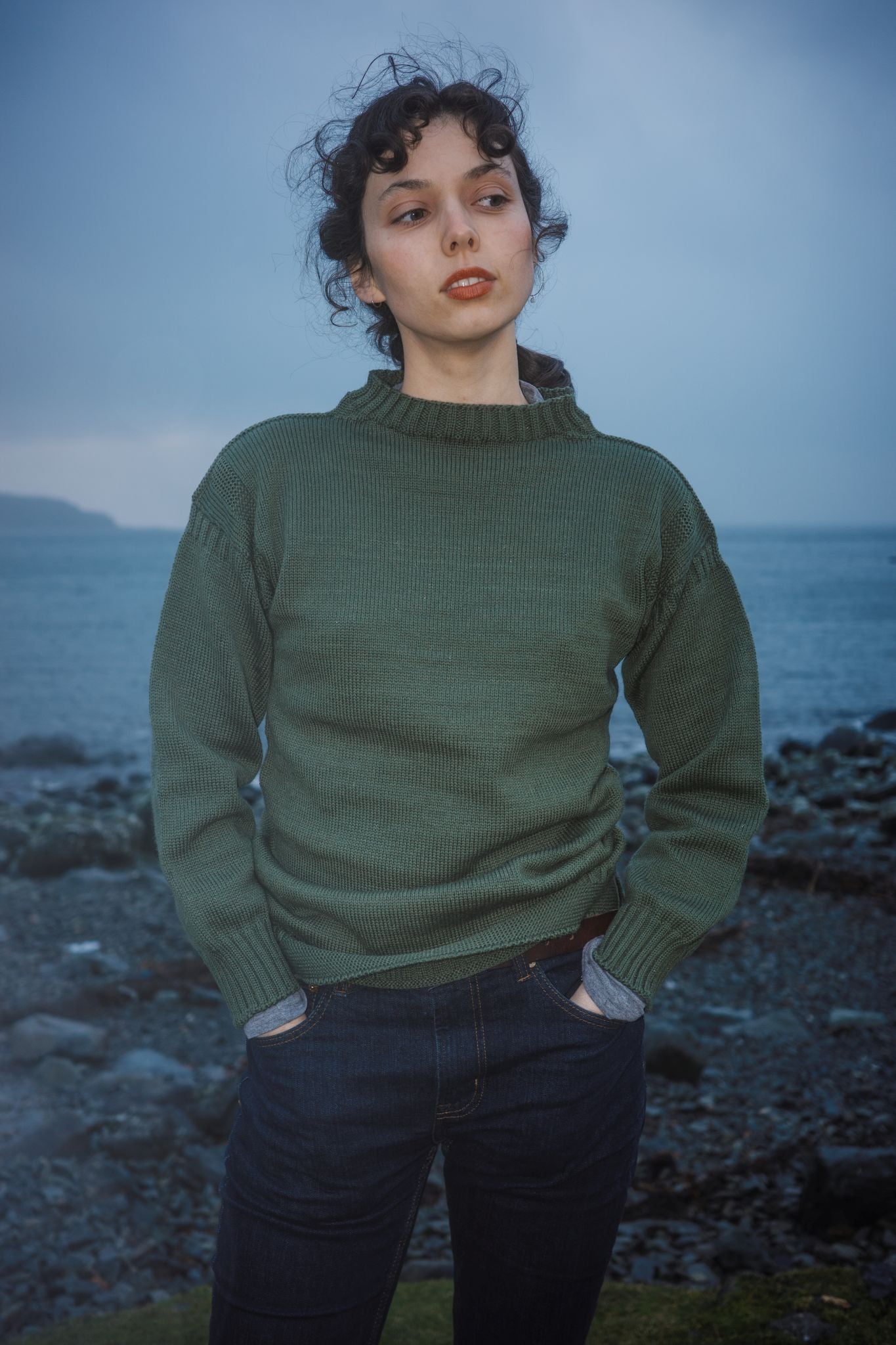 Female model wearing a sage green coloured traditional Guernsey abeside a lake with mountains behind 